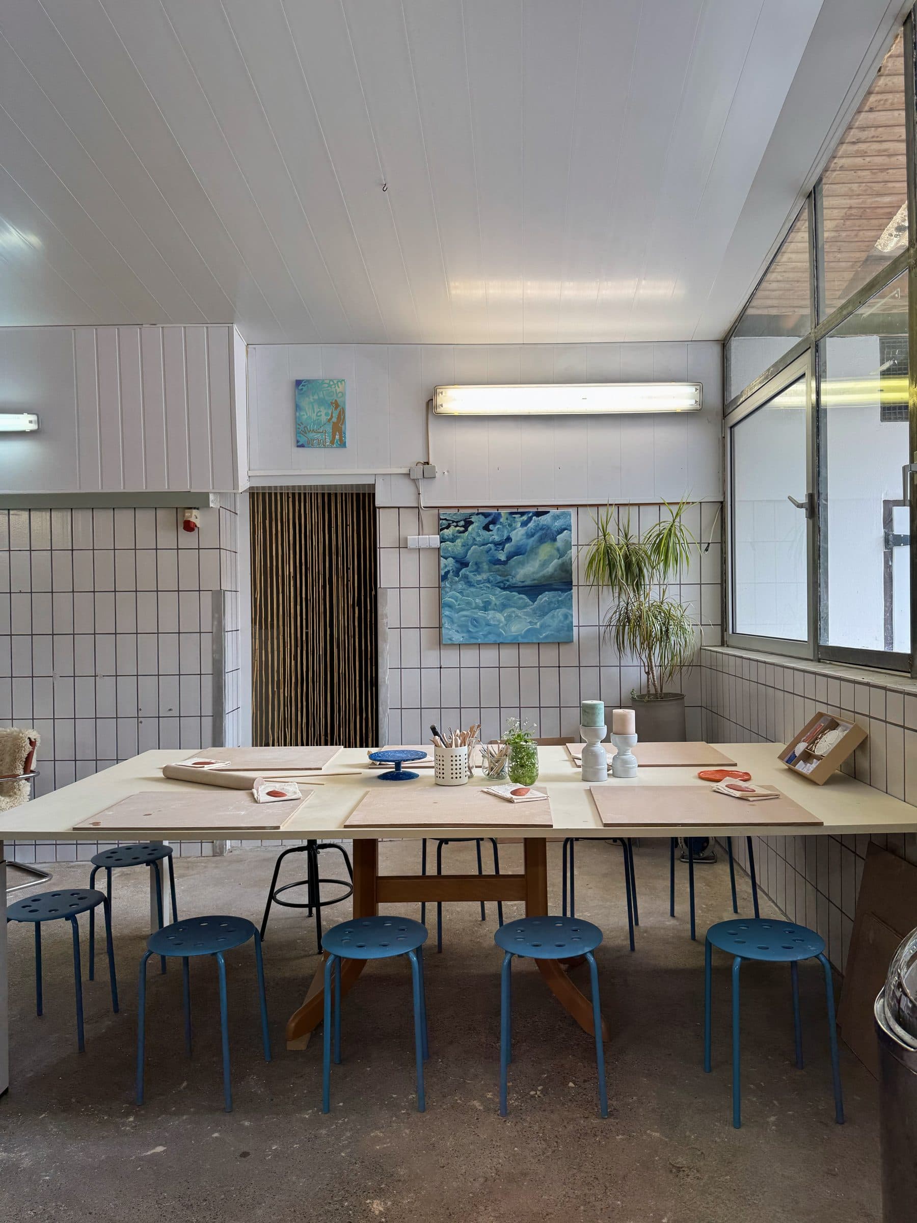 Heller Atelierraum mit Werktisch, blauen Hockern und Bildern an der Wand